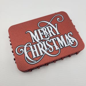 May include: A red, textured, rectangular sign with the words "Merry Christmas" in white cursive lettering with black outlines.