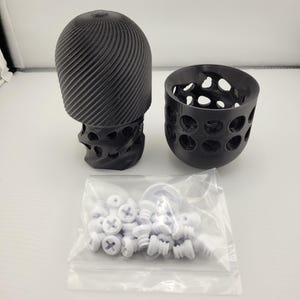 May include: A black 3D-printed sculpture with a skull-like base and a spiraling top, next to a perforated cup and a bag of white screws. The sculpture has a unique, textured design. The objects are on a white surface.