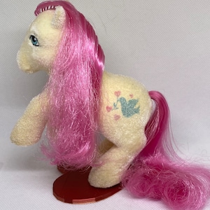 May include: A vintage My Little Pony toy with a cream-coloured body, pink mane and tail, and a blue bird with hearts design on its flank. The pony is standing on a red heart-shaped base.