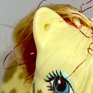 Puede incluir: Un juguete My Little Pony amarillo con un ojo azul, pesta&ntilde;as negras y una crin rosa y dorada. El pony tiene una mancha blanca en la cara y una cinta rosa y dorada en su crin.