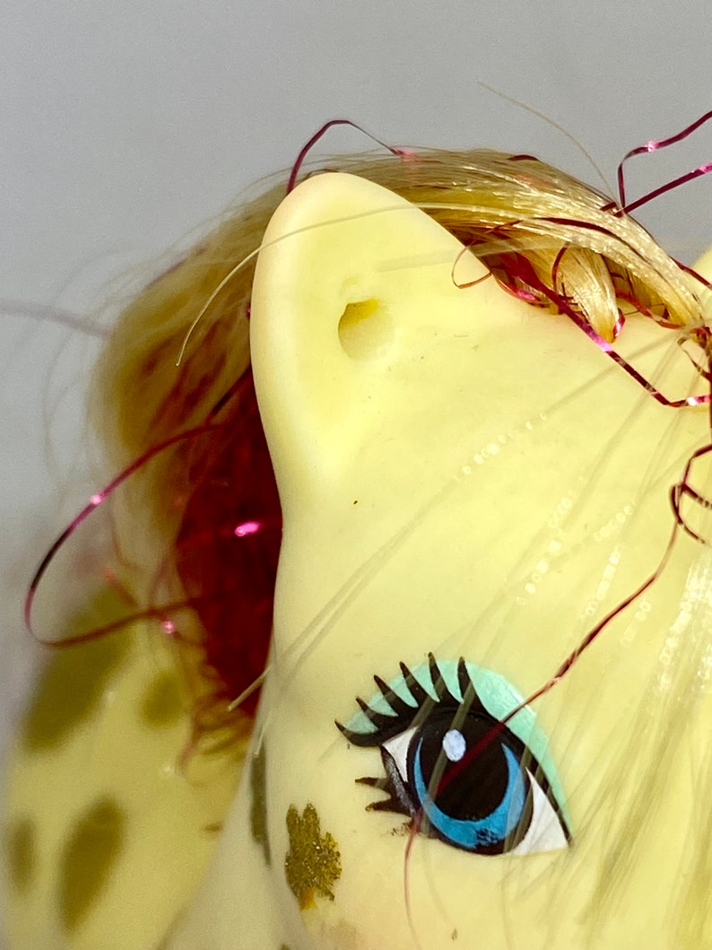 Puede incluir: Un juguete My Little Pony amarillo con un ojo azul, pesta&ntilde;as negras y una crin rosa y dorada. El pony tiene una mancha blanca en la cara y una cinta rosa y dorada en su crin.