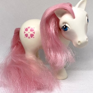 May include: A white pony with pink hair and a pink and white symbol on its flank. The pony has blue eyes and a white body.
