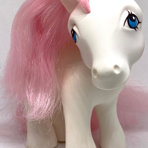 May include: A white pony toy with pink hair and blue eyes. The pony is standing on a white surface.