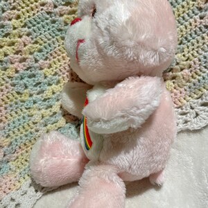 Puede incluir: Oso de peluche rosa con un arco&iacute;ris en el pecho. El oso est&aacute; sentado en una manta de ganchillo.