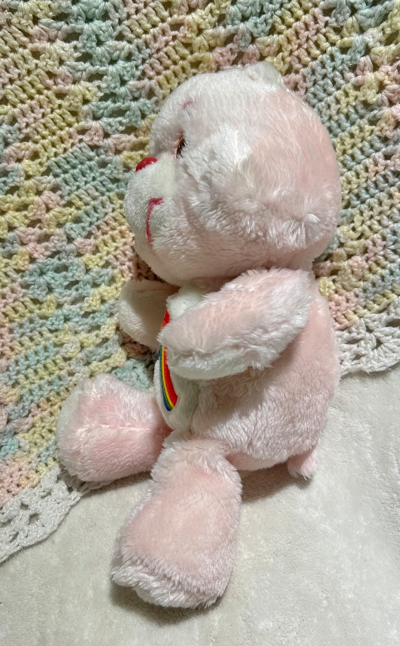 Puede incluir: Oso de peluche rosa con un arco&iacute;ris en el pecho. El oso est&aacute; sentado en una manta de ganchillo.