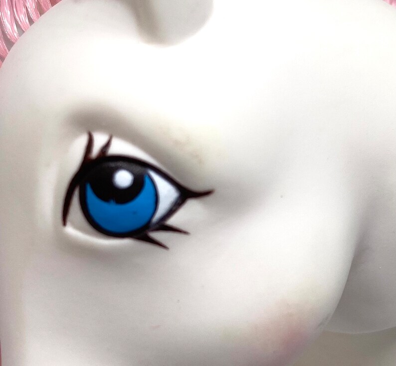 May include: Close-up of a blue eye with black eyelashes and a white pupil on a white plastic toy horse.