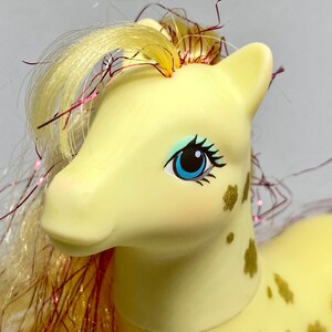 Puede incluir: Un pony amarillo con una larga melena y cola rubias con purpurina rosa y dorada. El pony tiene ojos azules y manchas doradas en el cuerpo.
