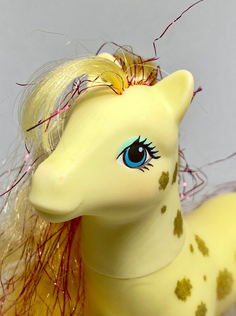 Puede incluir: Un pony amarillo con una larga melena y cola rubias con purpurina rosa y dorada. El pony tiene ojos azules y manchas doradas en el cuerpo.