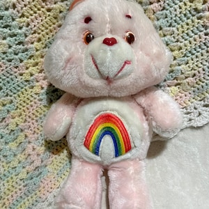 Puede incluir: Oso de peluche rosa con un arco&iacute;ris en el pecho. El oso tiene una nariz de coraz&oacute;n rojo y ojos marrones.