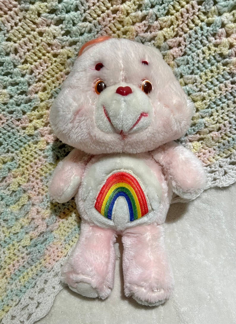 Puede incluir: Oso de peluche rosa con un arco&iacute;ris en el pecho. El oso tiene una nariz de coraz&oacute;n rojo y ojos marrones.