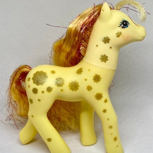 Puede incluir: Un juguete My Little Pony amarillo con un dise&ntilde;o de purpurina dorada y una larga melena rubia con rayas rojas.