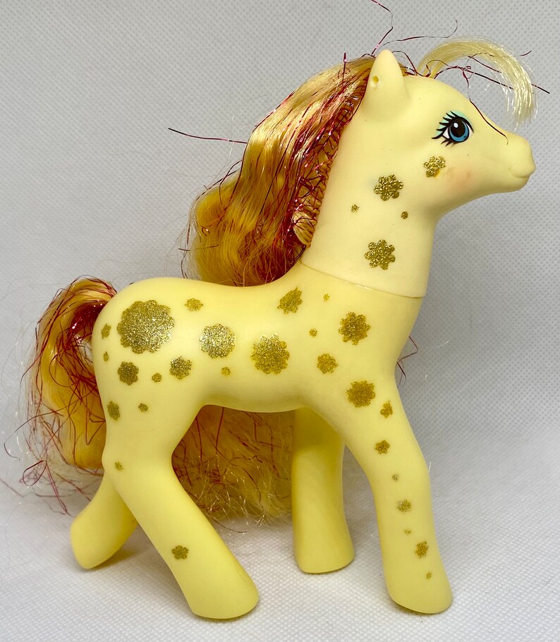 Puede incluir: Un juguete My Little Pony amarillo con un dise&ntilde;o de purpurina dorada y una larga melena rubia con rayas rojas.