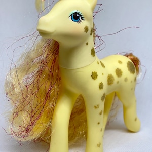 Puede incluir: Un juguete de pony amarillo con un patr&oacute;n de purpurina dorada y una larga melena fluida con purpurina roja y dorada. El pony tiene ojos azules y una nariz rosa.