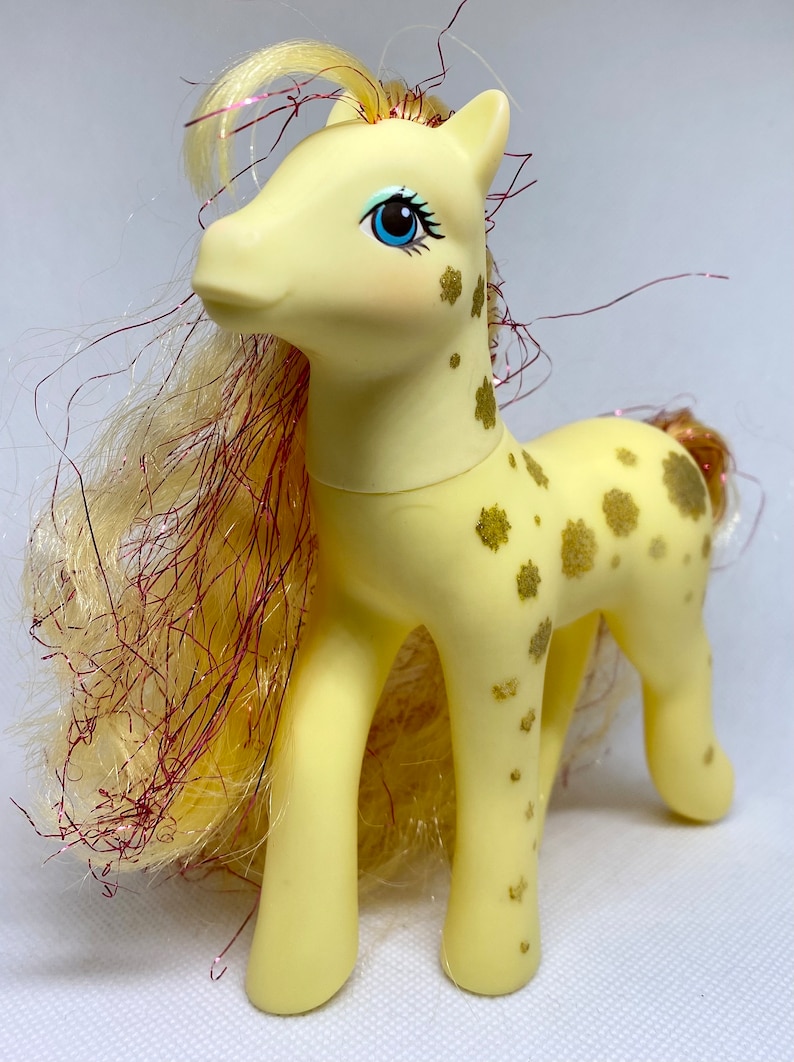 Puede incluir: Un juguete de pony amarillo con un patr&oacute;n de purpurina dorada y una larga melena fluida con purpurina roja y dorada. El pony tiene ojos azules y una nariz rosa.