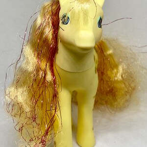 Puede incluir: Un juguete My Little Pony amarillo con una melena y cola rosa y dorada. El pony tiene ojos azules y un cuerpo amarillo con manchas marrones.