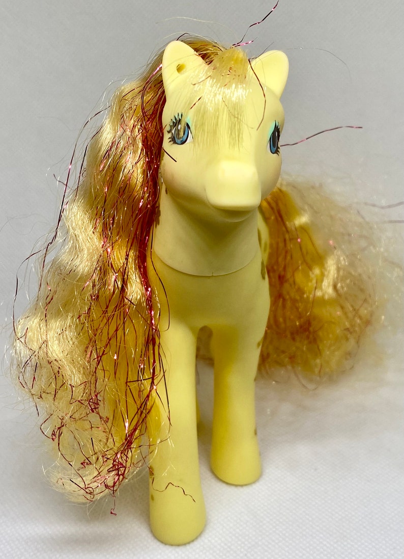 Puede incluir: Un juguete My Little Pony amarillo con una melena y cola rosa y dorada. El pony tiene ojos azules y un cuerpo amarillo con manchas marrones.