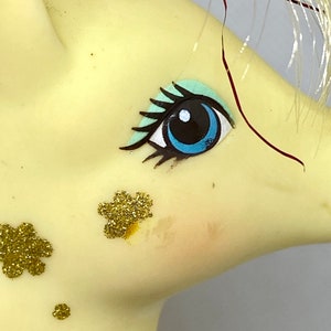 Puede incluir: Primer plano de la cara de un pony de juguete amarillo con ojos pintados de azul y verde y detalles de purpurina dorada.