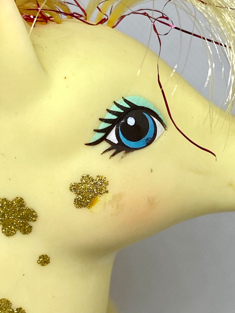 Puede incluir: Primer plano de la cara de un pony de juguete amarillo con ojos pintados de azul y verde y detalles de purpurina dorada.