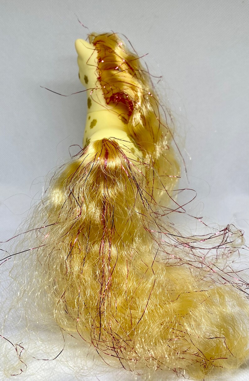 Puede incluir: Un caballo de juguete amarillo con una melena y una cola largas y fluidas. La melena y la cola est&aacute;n hechas de un material suave y esponjoso y tienen un brillo rosado. El caballo tiene manchas marrones en su cuerpo.