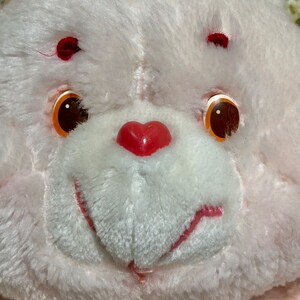 Puede incluir: Primer plano de un oso de peluche blanco con una nariz roja en forma de coraz&oacute;n y ojos naranjas. El oso tiene un rubor rosado en las mejillas.