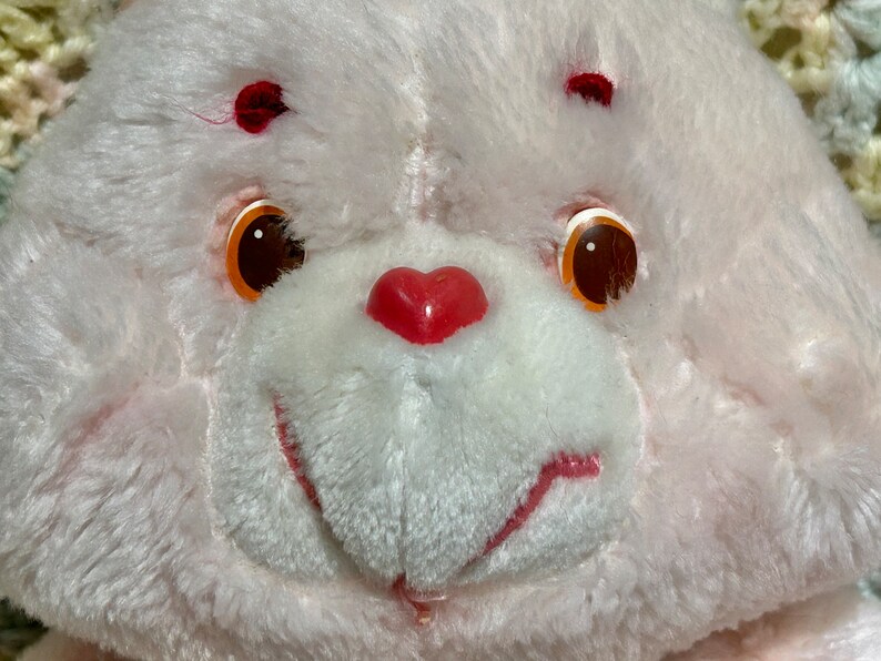 Puede incluir: Primer plano de un oso de peluche blanco con una nariz roja en forma de coraz&oacute;n y ojos naranjas. El oso tiene un rubor rosado en las mejillas.