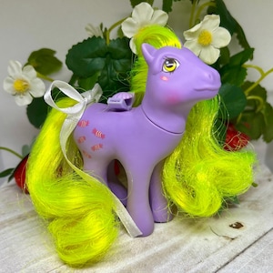 May include: A purple plastic pony figurine with bright neon yellow hair and a white ribbon tied in its mane. The pony features a cutie mark of several small pink candies on its flank, pink cheek blush, and large yellow eyes with black pupils.