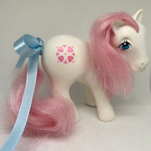 May include: White pony with pink hair and a blue bow. The pony has a pink flower symbol on its flank.