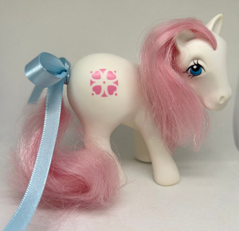 May include: White pony with pink hair and a blue bow. The pony has a pink flower symbol on its flank.