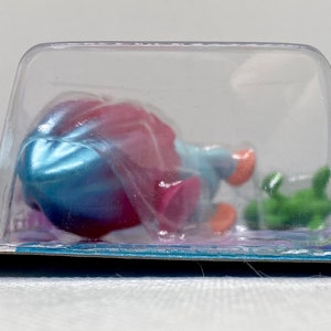 Puede incluir: Una figura de sirena de juguete de pl&aacute;stico en una concha azul y rosa con un caballito de mar verde. La figura est&aacute; empaquetada en una concha de pl&aacute;stico transparente.