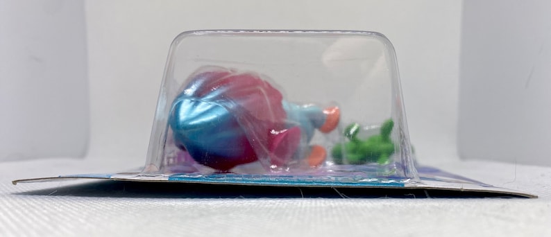 Puede incluir: Una figura de sirena de juguete de pl&aacute;stico en una concha azul y rosa con un caballito de mar verde. La figura est&aacute; empaquetada en una concha de pl&aacute;stico transparente.