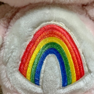 Puede incluir: Juguete de peluche rosa con un arco&iacute;ris blanco bordado en su vientre.