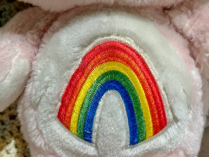 Puede incluir: Juguete de peluche rosa con un arco&iacute;ris blanco bordado en su vientre.