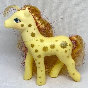 Puede incluir: Un pony My Little Pony amarillo con un patr&oacute;n de flores de purpurina dorada. El pony tiene una melena y una cola largas y fluidas con reflejos rojos y dorados.