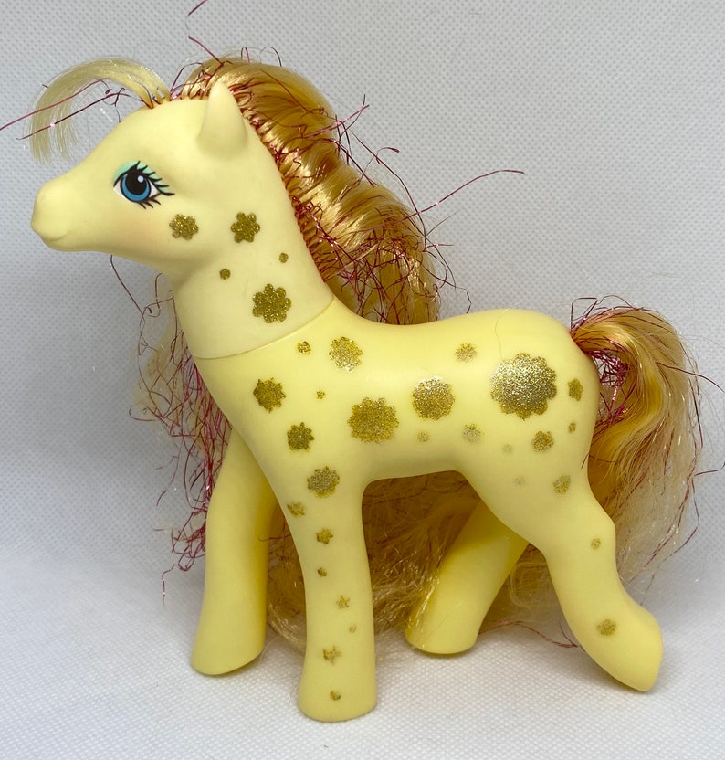Puede incluir: Un pony My Little Pony amarillo con un patr&oacute;n de flores de purpurina dorada. El pony tiene una melena y una cola largas y fluidas con reflejos rojos y dorados.