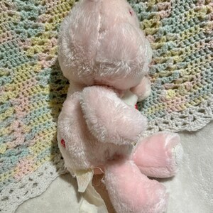 Puede incluir: Oso de peluche rosa con un coraz&oacute;n rojo en el pecho. El oso est&aacute; sentado en una manta de crochet blanca.