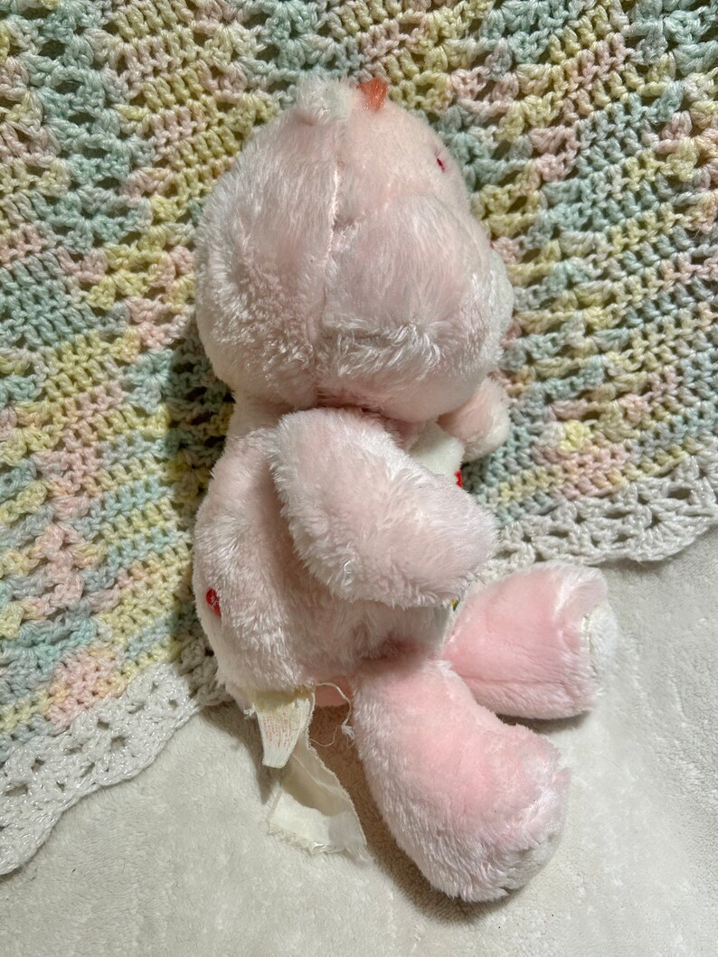 Puede incluir: Oso de peluche rosa con un coraz&oacute;n rojo en el pecho. El oso est&aacute; sentado en una manta de crochet blanca.