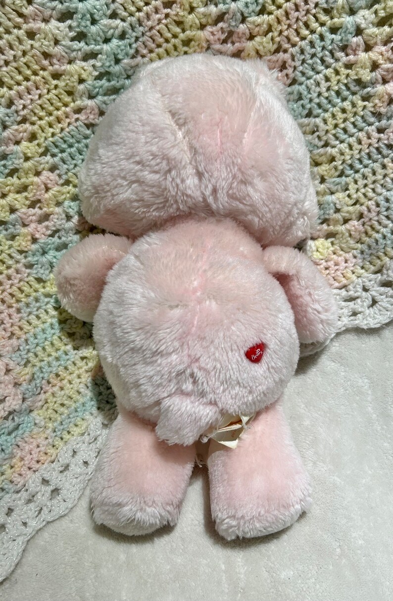 Puede incluir: Oso de peluche rosa con un coraz&oacute;n rojo en el vientre. El oso est&aacute; sentado en una manta de ganchillo.