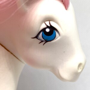 May include: Close-up of a white pony toy with a pink mane and tail. The pony has a blue eye with black eyelashes and a black pupil.