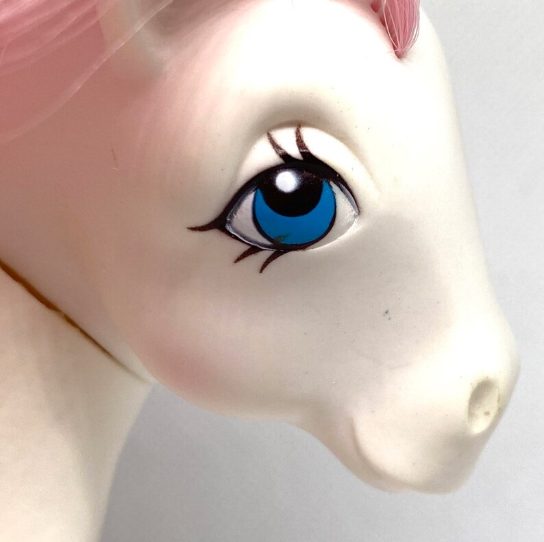 May include: Close-up of a white pony toy with a pink mane and tail. The pony has a blue eye with black eyelashes and a black pupil.