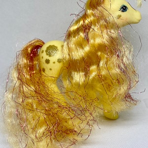 Puede incluir: Un pony My Little Pony amarillo con un patr&oacute;n de purpurina dorada y una larga melena amarilla y rosa.