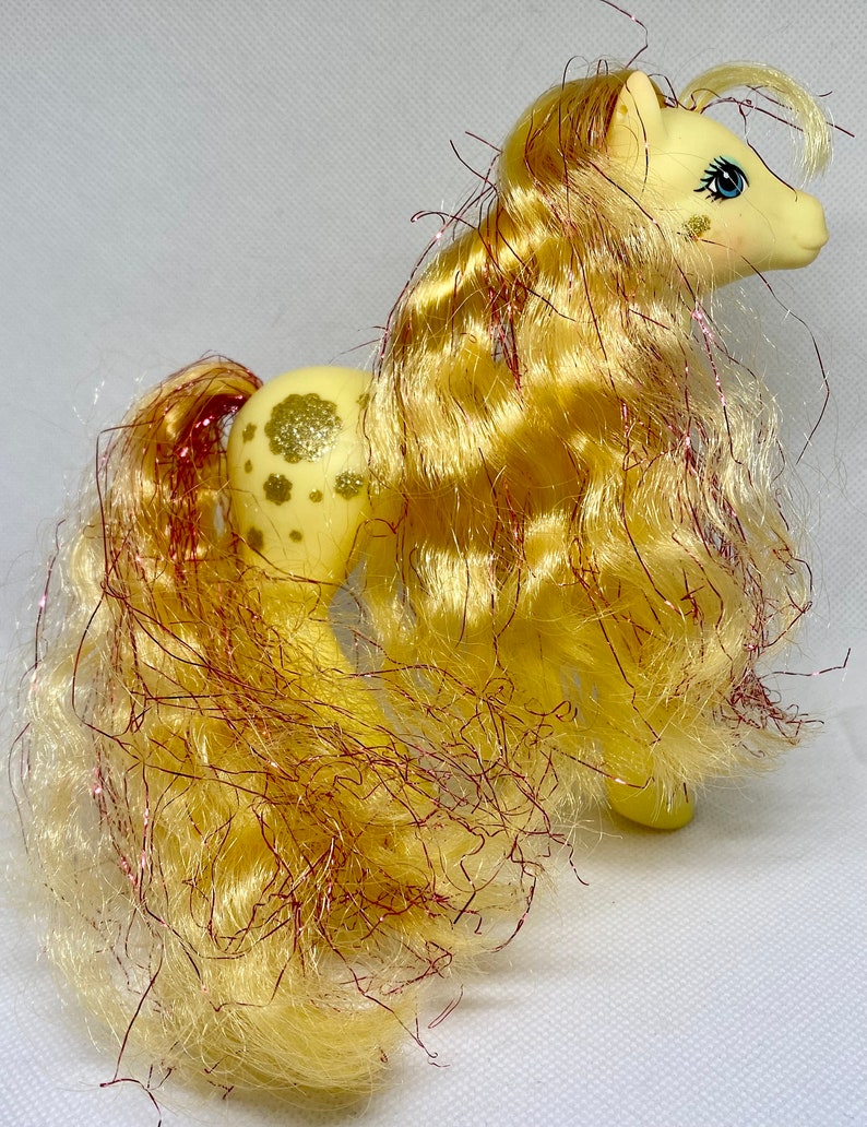 Puede incluir: Un pony My Little Pony amarillo con un patr&oacute;n de purpurina dorada y una larga melena amarilla y rosa.