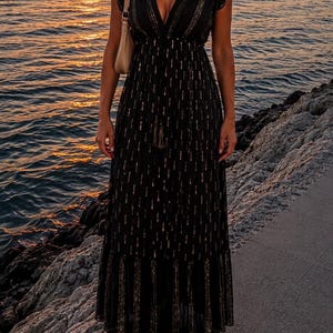 Peut inclure: Une robe longue noire dotée d'un décolleté en V profond, de manches courtes volantées et d'une taille cintrée avec un cordon à pompons. Le tissu est orné de rayures métalliques dorées verticales et d'un ourlet à volants, avec un sac à épaule beige.