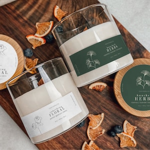 Könnte beinhalten: Zwei Glaskerzenbehälter mit Holzdeckeln. Die Kerzen sind mit "something floral" mit dem Duft "Jasmin, Geranie und Poesie" und "bunched herbs" mit dem Duft "Rosmarin, Lavendel und Zitronengras" beschriftet.