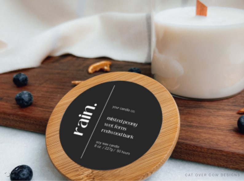 Single candle and candle lid on kitchen counter. Candle as a black label with luxury serif font written on the left. Asymmetric design for editable text of the candle company, scents, volume to the right of the candle name separated by bold line