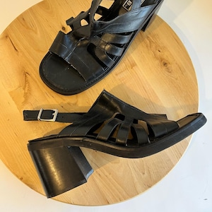 May include: A pair of black leather sandals with a cage-like design. The sandals have a square toe, a thick block heel, and an adjustable ankle strap with a silver buckle. The heels are approximately 3 inches tall.