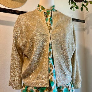 May include: A vintage gold sequin jacket with three-quarter sleeves is displayed on a mannequin. The jacket is paired with a floral print dress in shades of green, blue, and cream. A straw hat hangs in the background.