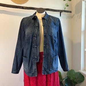 May include: A dark blue denim jacket with a textured appearance, featuring a collared neckline, three front pockets, and button closures. The jacket is displayed on a mannequin, paired with a red skirt. The jacket's design includes long sleeves.