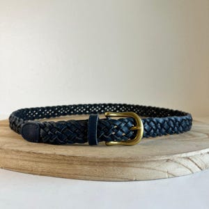 May include: A navy blue braided belt with a gold-colored buckle. The belt is made of woven leather and is displayed on a wooden surface. The belt is a classic accessory.