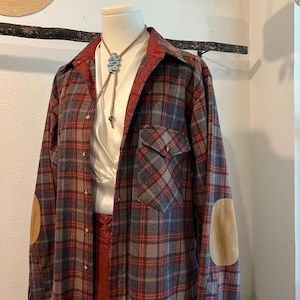 Może przedstawiać: Koszula w kratę w kolorze czerwonym, niebieskim i brązowym, z zamszowymi łokciami i czerwonym kołnierzem. Widoczna jest biała bluzka i krawat bolo z turkusowym wzorem. Koszula jest prezentowana na manekinie.