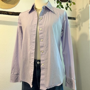 Könnte beinhalten: Ein hellviolettes Langarmhemd mit Knopfleiste, klassischem Kragen und weißen Knöpfen, präsentiert auf einer Schaufensterpuppe. Das Hemd verfügt über eine strukturierte Passform mit durchgehender Knopfleiste, langen Ärmeln mit Manschetten und glattem Stoff.